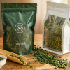 Tea packaging bags on a wooden table, a matte green aluminium foil stand-up pouch and a clear high-barrier pouch holding green tea leaves with a ceramic cup and bamboo scoop