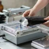 Tabletop impulse heat sealer closing a matte black stand-up coffee pouch on a stainless steel packing bench
