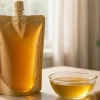 Matte amber stand-up pouch with a screw spout filled with golden honey beside a glass dish of honey and a wooden dipper on a light wooden table