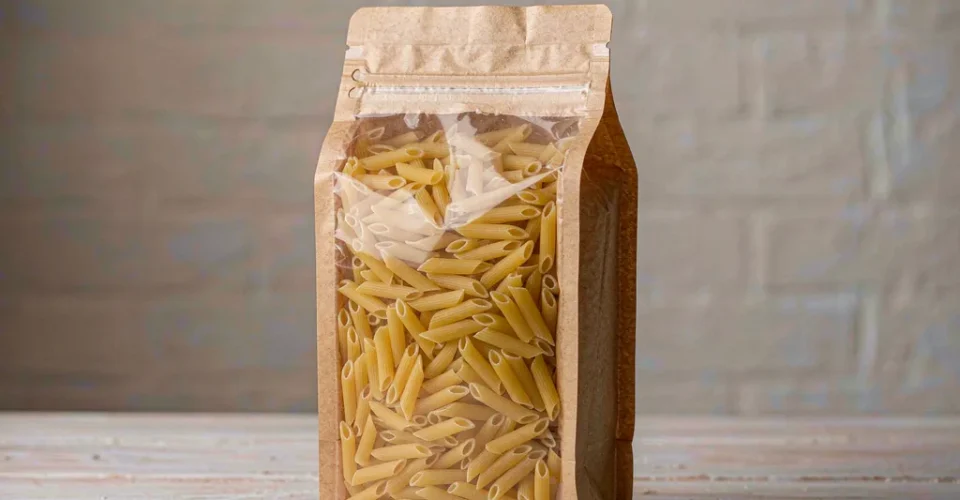 Kraft-front stand-up pasta packaging bag with a clear window and zipper, filled with dried penne on a wooden table